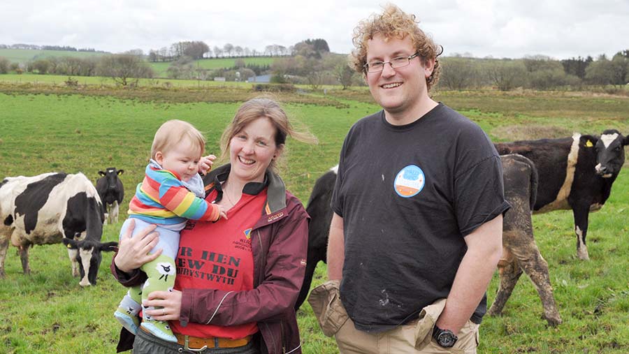 Liam and Annie James stand in a field of dairy cows. Annie holds infant daughter Buddug