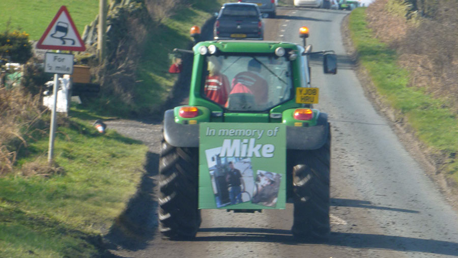 A tractor travels along a road with a sign which reads: In memory of Mike"