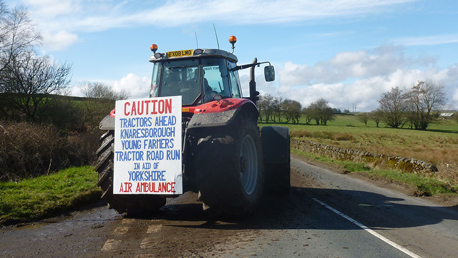 A tractor stands in the road. It has a sign attached. The sign reads: "Caution tractors ahead. Knaresborough young farmers' tractor road run in aid of Yorkshire air ambulance."