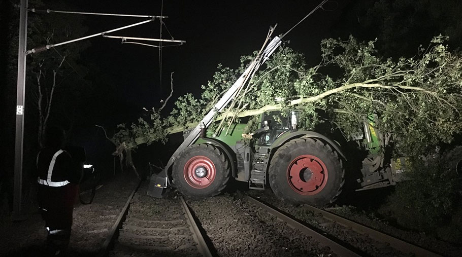 A tractor and trailer on railways tracks. An uprooted tree lies across the cab