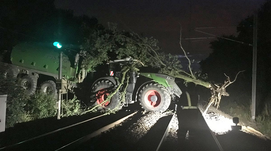 A tractor and trailer on railway tracks. An uprooted tree lies across the cab