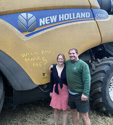 Sally and John standing next to New Holland with "Will you marry me" written in the dust