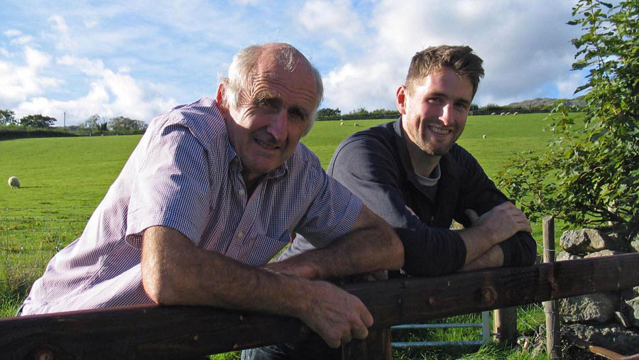 A father-and-son pair of farmers lean against a fence from within a field