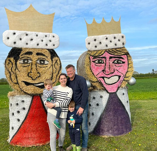 Farming couple posing with two children in front of bales featuring King Charles and Queen Camilla designs