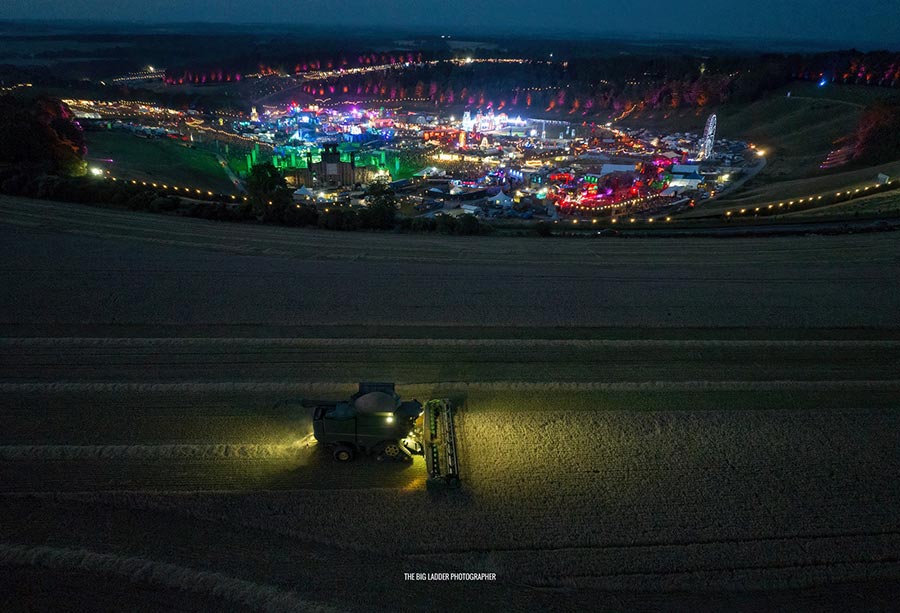 Aerial view of a combine harvester cutting spring barley at night as a lights from a lively music festival shine in the other field