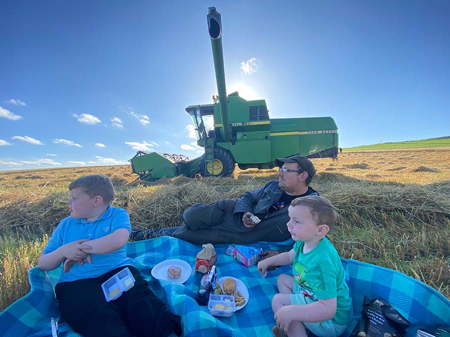 Man and two children eating a picnic on the grass with a combine in view in the background