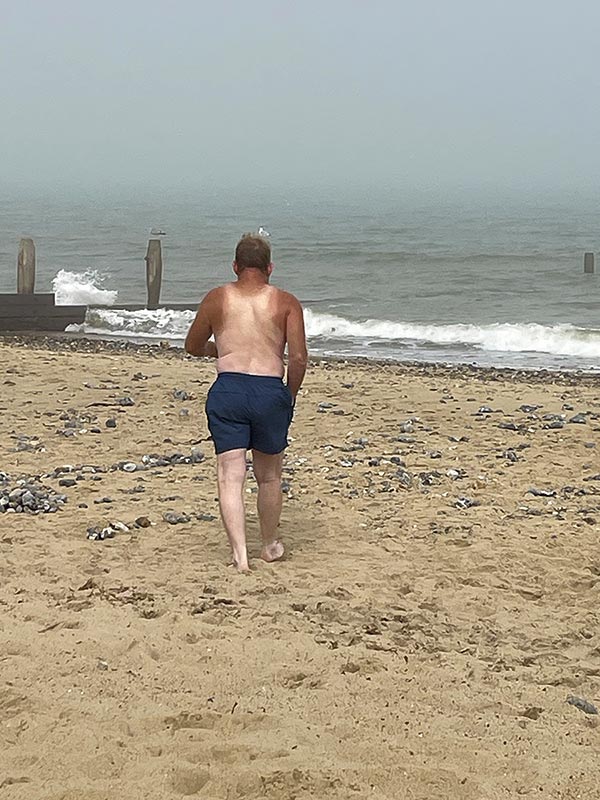 View of David Cross walking into the sea with tanned arms and neck and a white back