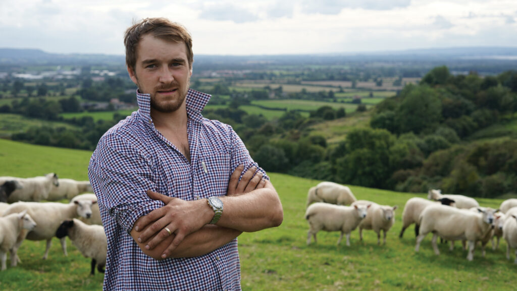 A man standing in a field with sheep behind him