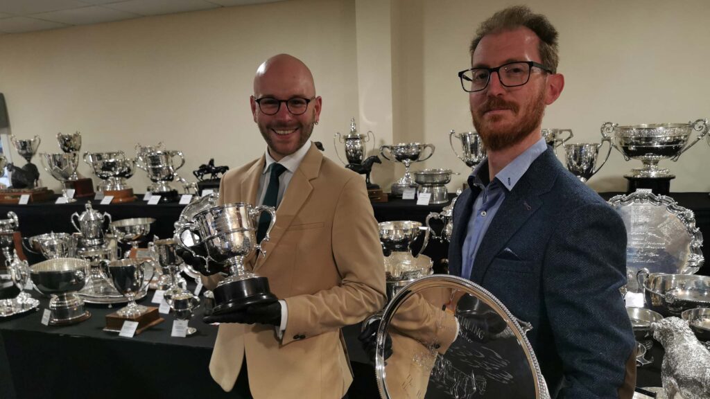 Two men in a room full of silver cups and trophies