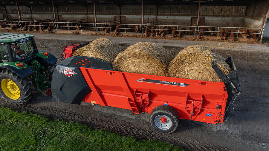 Green tractor pulling red Kuhn Primor 3570 bale processor loaded with hay bales near a cattle barn.