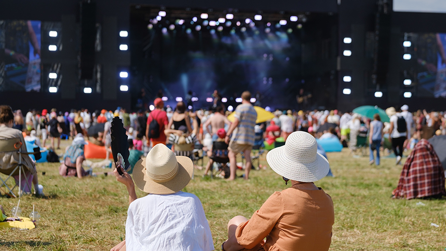 Outdoor music festival scene with a crowd in front of a brightly lit stage, performers in the background, and two people in wide-brimmed hats seated on the grass in the foreground, one holding a fan.