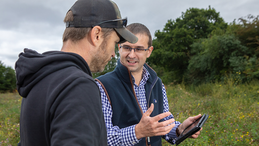 Two people standing in a field, with one holding a tablet and gesturing while talking to the other.