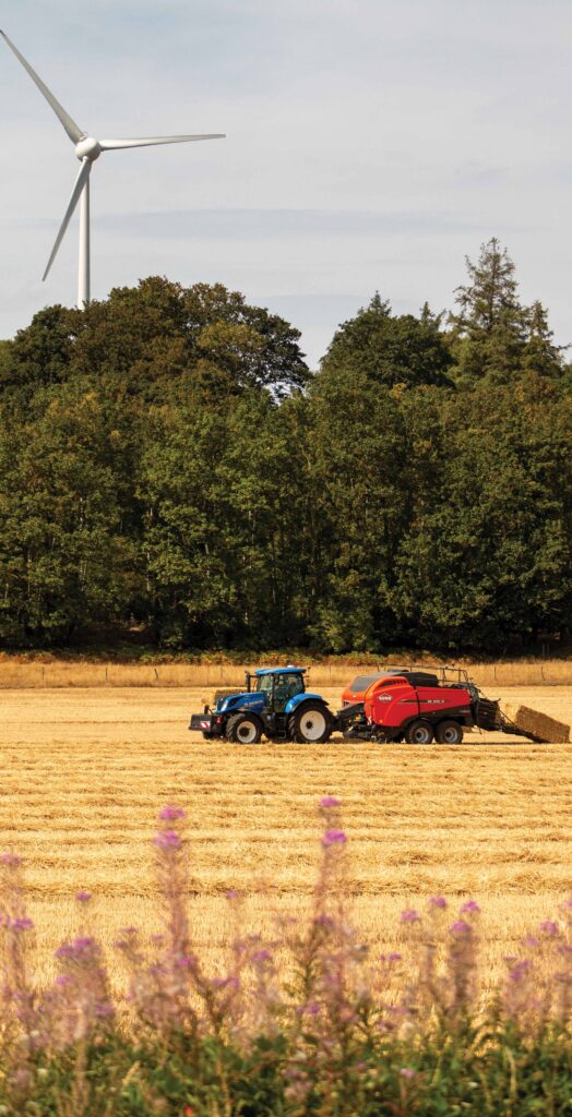 A New Holland T7.245 AutoCommand tractor and Kuhn SB 1290 ID baling wheat straw, with a pretty pink floristic hedgerow in the foreground and wind turbine in the background