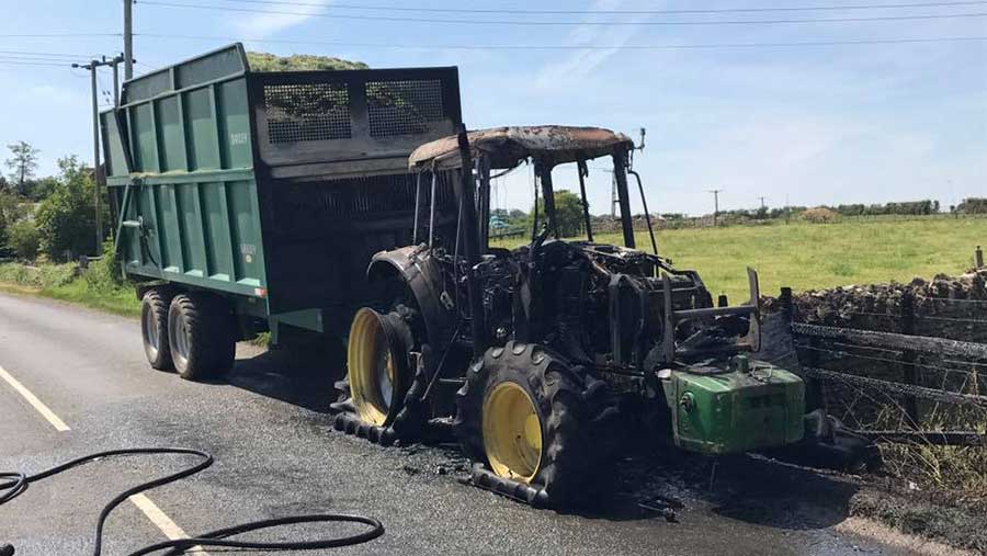 A burned out tractor stands in a road. Behind it is a trailer filled with silage