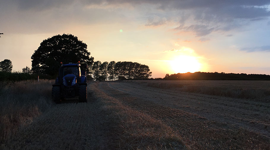 A tractor pulling a trailer moves through a field as the sun sets behind it