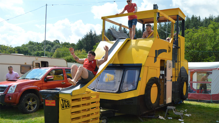 A caravan is decked out in JCB yellow. A rooftop slide has been added. Men watch as one of their group uses the slide