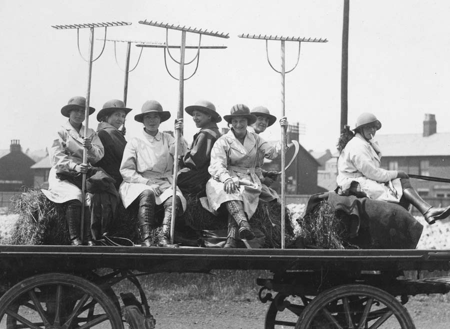 A black-and-white image shows a group of women riding on trailer holding farm kit