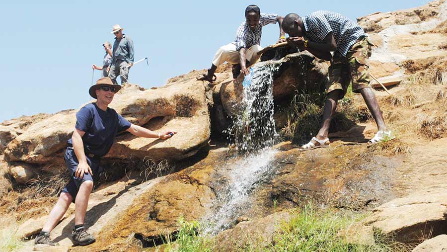 Farmer Richard Gibson stands on a rocky cliff face while two Kenyan men stand to his right next to water rushing down a slope