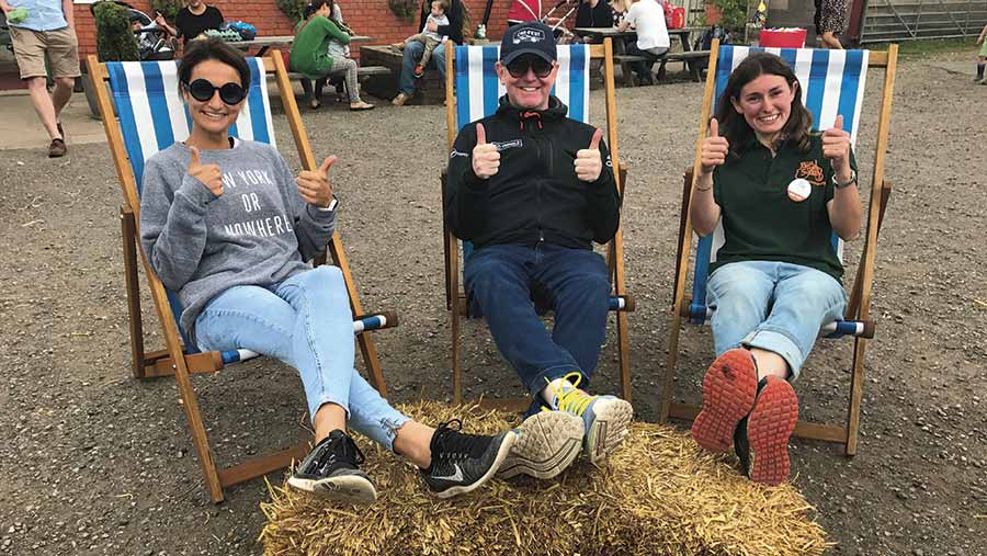 Radio 2 star Chris Evans sits on a deckchair with his feet on a straw bale. To his left is his wife Natasha Shishmanian and to his right is Alison Frost, Row Farm’s OFS organiser. All three are giving the photograph a thumbs up sign 